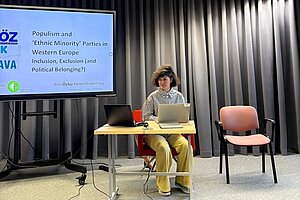 Öykü presenting research in Tallinn, dressed in a white and black shirt with yellow pants like a derivative version of the supervillain the Riddler.