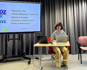 Öykü presenting research in Tallinn, dressed in a white and black shirt with yellow pants like a derivative version of the supervillain the Riddler.